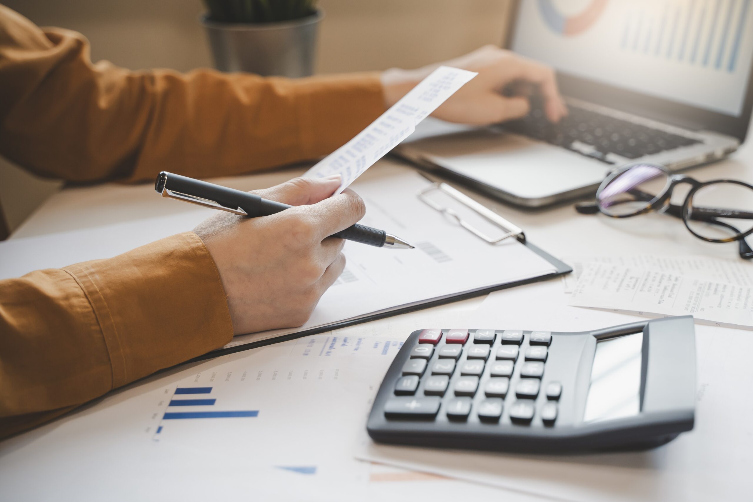 desk with paperwork, calculator and a pair of hands tapping on the laptop